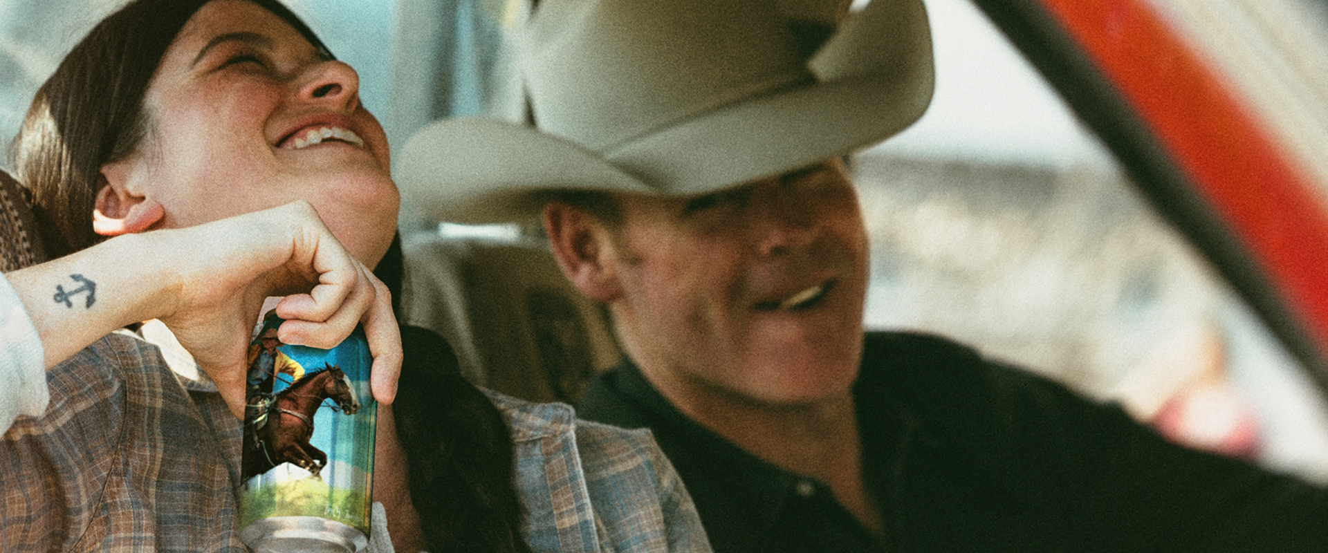 Smiling woman holding a drink can with a cowboy-on-horse illustration, seated beside a man in a cowboy hat in a truck.
