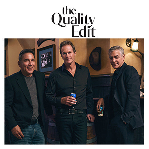 Three men standing in a dimly lit bar holding cans of beer, posed in front of wooden barrels and framed photos