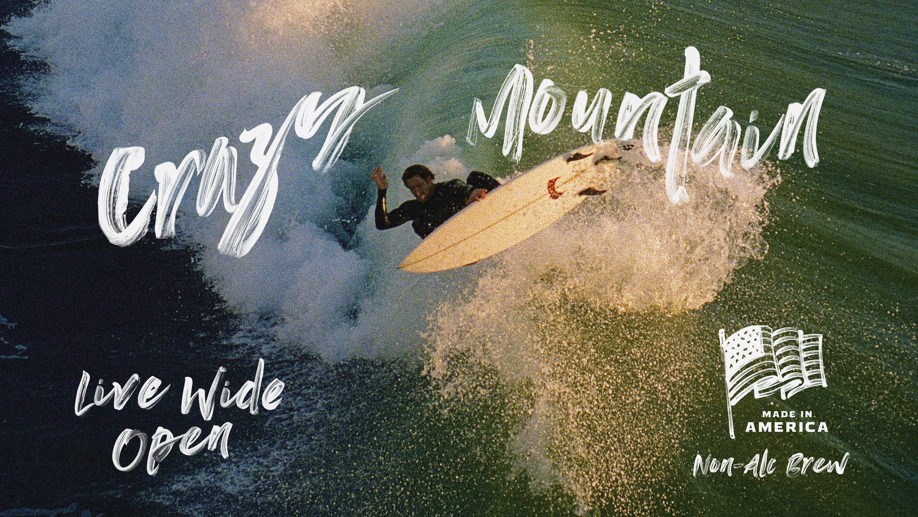 Man in a black wetsuit surfing a large wave with splashes of the wave consuming the image background. Words 'Crazy Mountain' appear in white brush strokes above his head, along with 'Live Wide Open' and 'Made in America Non-Alc Brew' below his surfboard and an American flag.