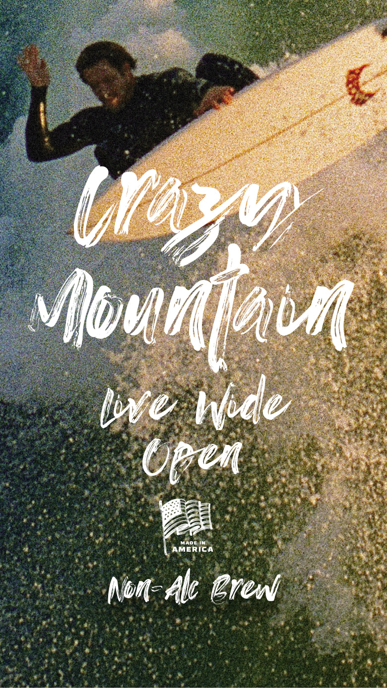Man in a black wetsuit surfing a large wave with splashes of the wave consuming the image background. Words 'Crazy Mountain' appear in white brush strokes along with 'Live Wide Open' and 'Made in America Non-Alc Brew' below his surfboard and an American flag. Mobile version of the image.
