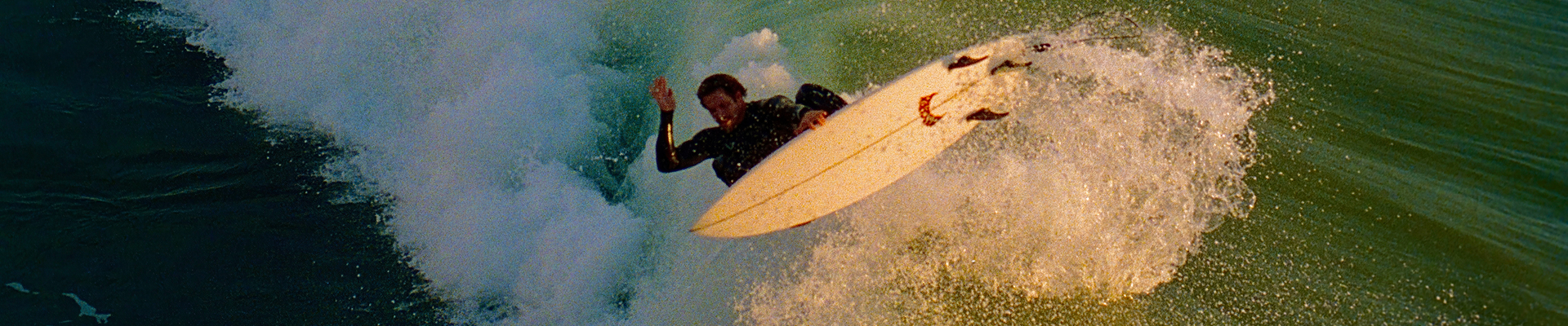 Surfer launching an aerial off a breaking green wave, white spray trailing behind the shortboard.