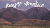 Cowboy rides horse across a mountain background, lassoing a rope. Behind the mountains shows 'Crazy Mountain' in white brushstrokes text. 